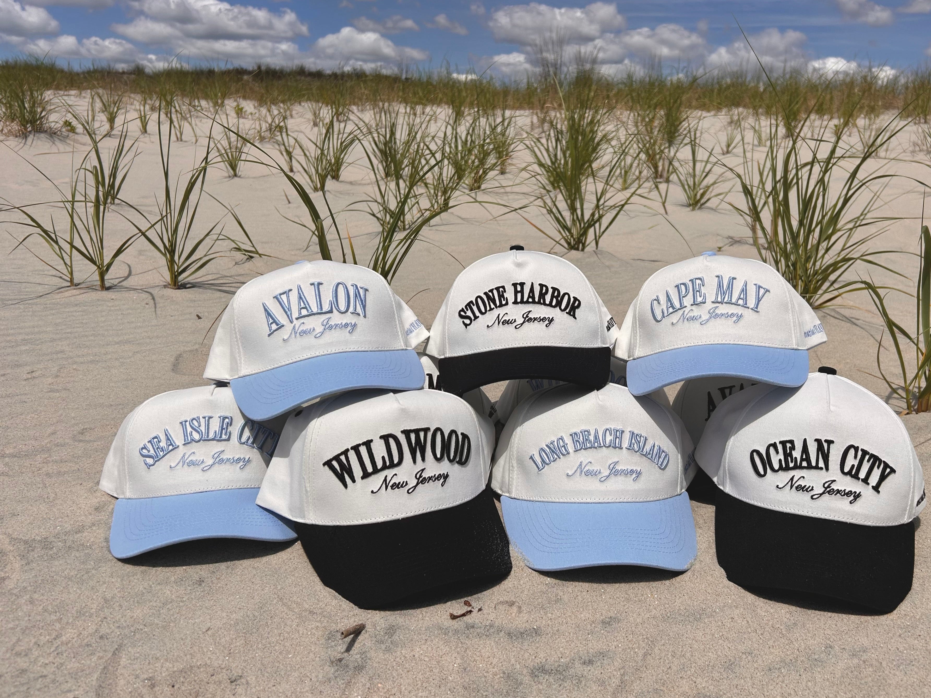 Collection of baseball caps with New Jersey locations on a sandy beach.